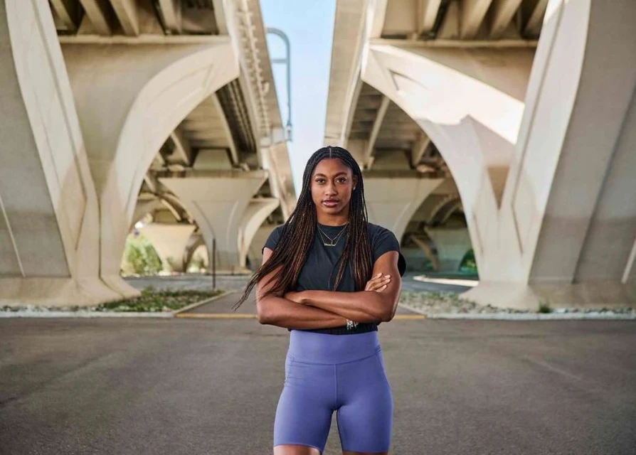 A confident Black woman poses with arms crossed under a concrete bridge. Wearing workout clothes, she stands on asphalt in neutral daylight.