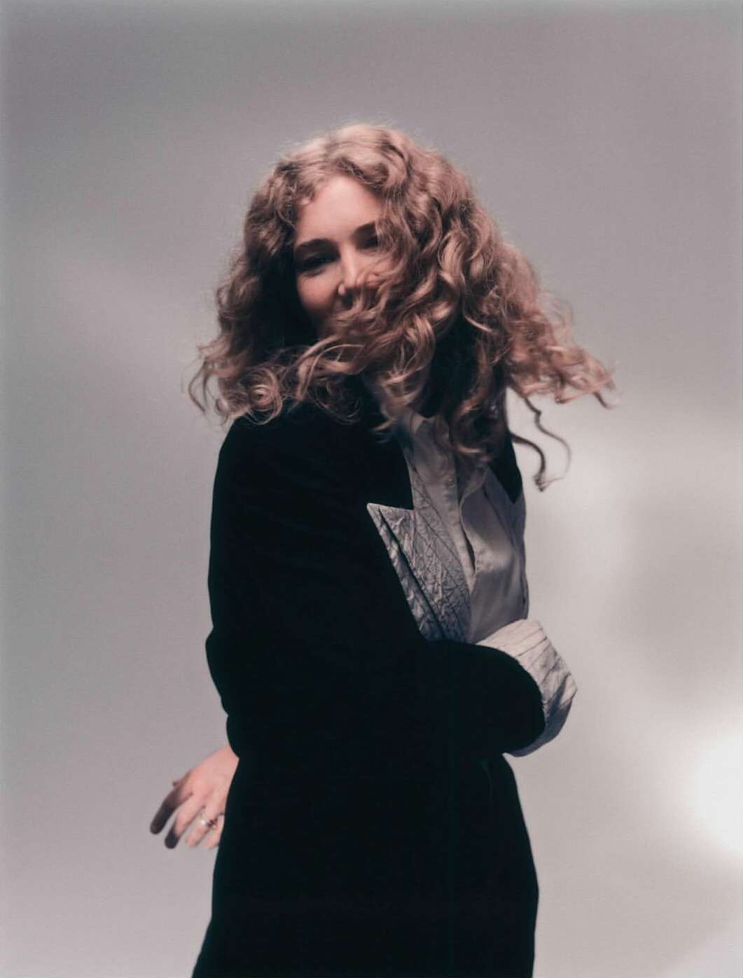Blonde woman in black jacket posing with curly hair against a neutral backdrop in a studio setting, fashionably dressed.