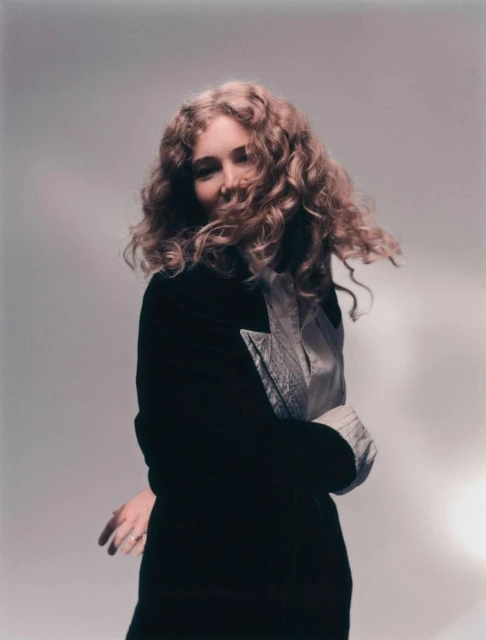 Blonde woman in black jacket posing with curly hair against a neutral backdrop in a studio setting, fashionably dressed.