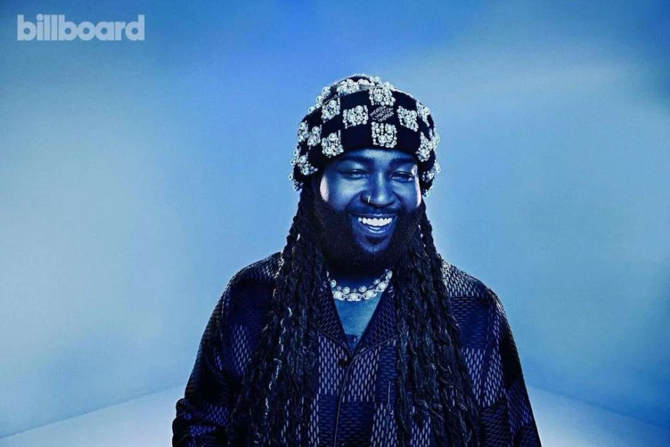 Close-up of a smiling adult male with long braids and a patterned beanie against a blue background, featuring the Billboard logo.