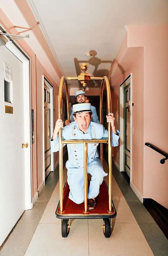 Two men in light blue uniforms ride a gold luggage cart in a hotel hallway with doors, pink walls, and grey striped carpet.