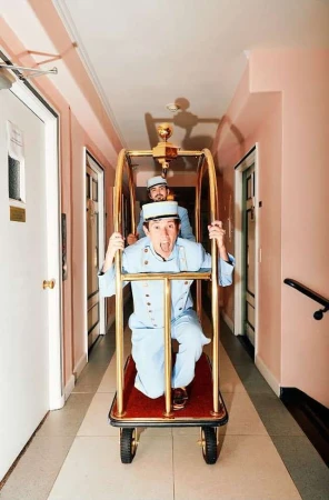 Two men in light blue uniforms ride a gold luggage cart in a hotel hallway with doors, pink walls, and grey striped carpet.