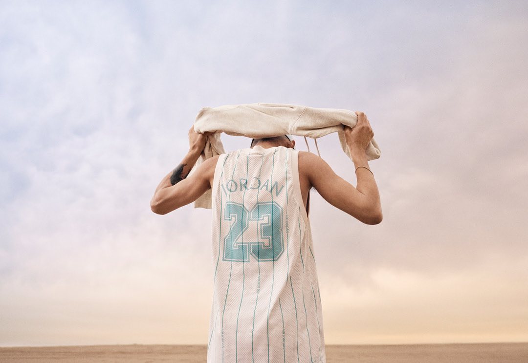 Back view of an adult in a sports jersey wiping their head with a towel, standing in a flat, sandy environment beneath a cloudy sky.