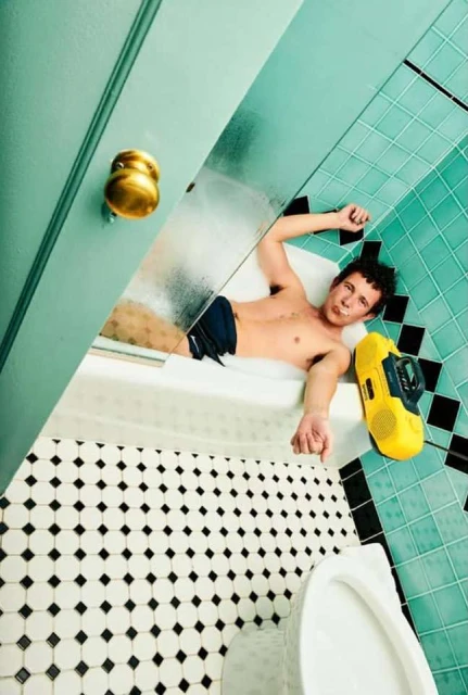 Man in bathtub with yellow boombox. Man is reclining and smiling.  Bathroom has turquoise tiled walls and black and white tiled floor.