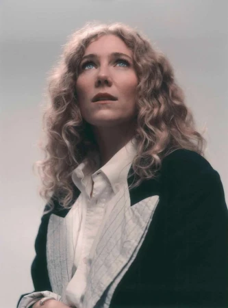 Portrait of a blonde woman with curly hair wearing a dark blazer and white pinstripe shirt looking upward against a pale background