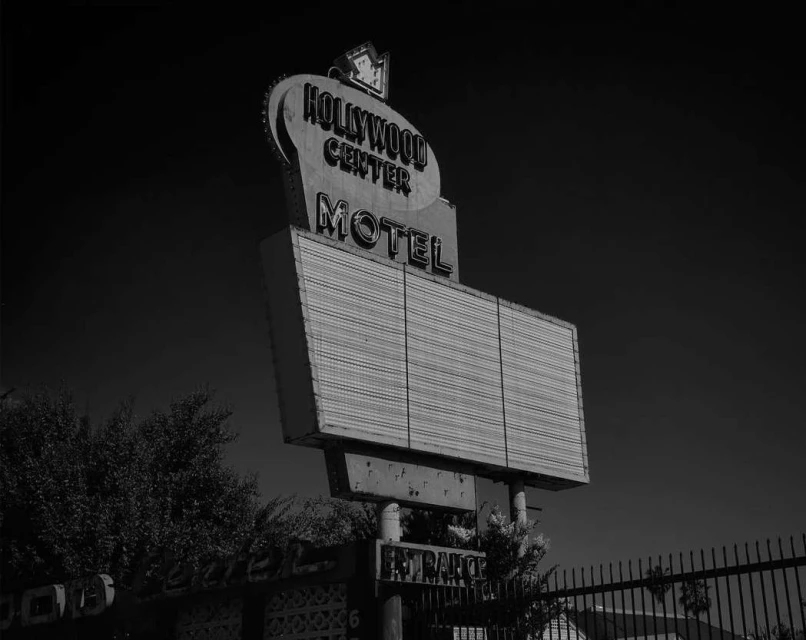 Black and white photo of the Hollywood Center Motel sign with an empty marquee, framed by trees and set against a dark sky.