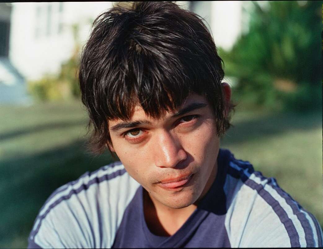 Portrait of a young adult male with dark hair wearing a blue and white striped shirt, looking directly at the camera in an outdoor setting.