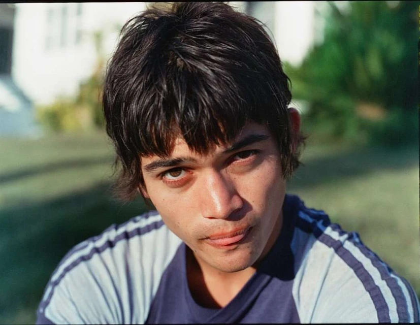 Portrait of a young adult male with dark hair wearing a blue and white striped shirt, looking directly at the camera in an outdoor setting.