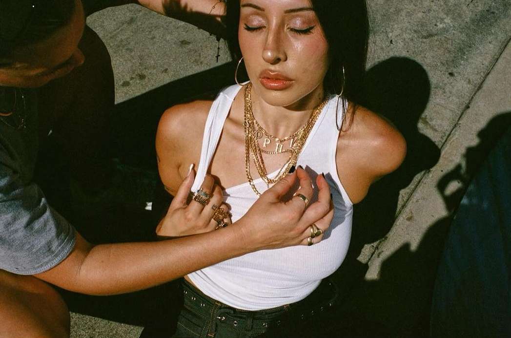 Close-up of a young adult woman with multiple gold chain necklaces posing with closed eyes, assisted by another person's hands on her white top.