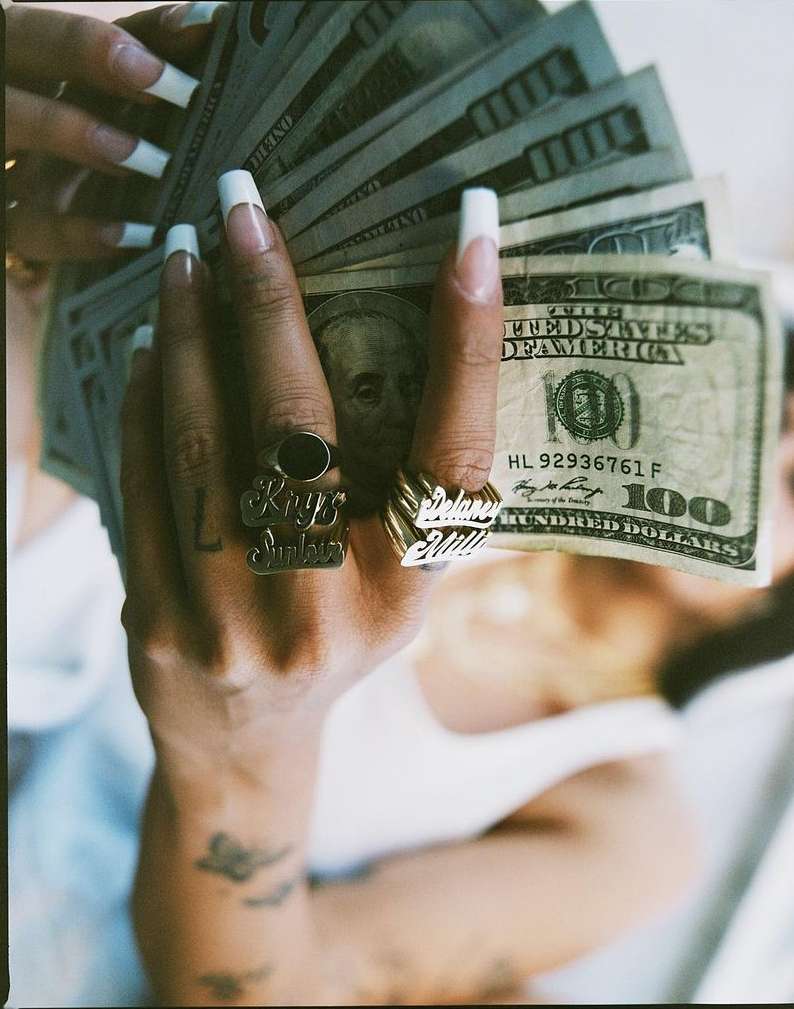 Close-up shot of a person's hand holding a fan of US hundred dollar bills, featuring ornate rings on the fingers and white-painted nails.