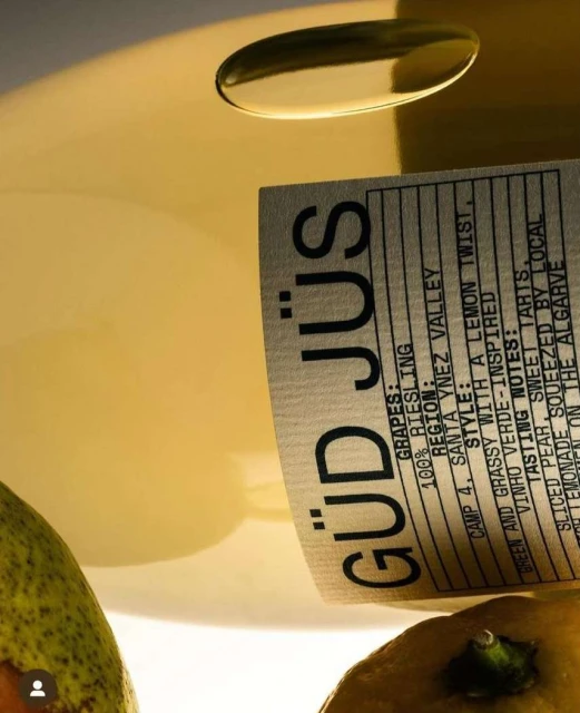 Close-up of a bottle of GÜD JÜS wine surrounded by pears and a light yellow background. The label is visible with bold, black font.