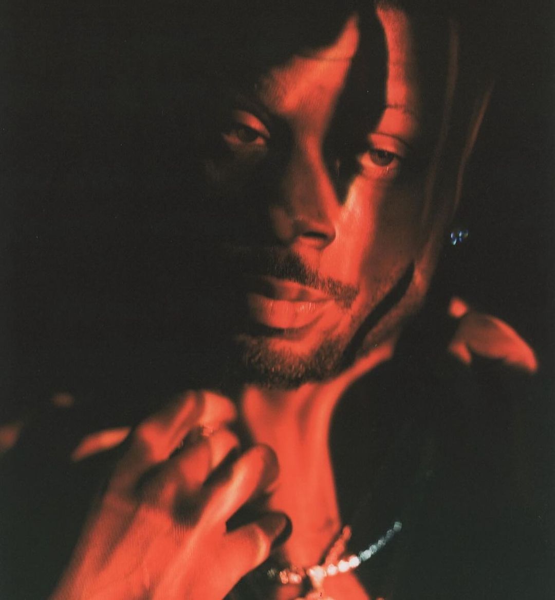 Portrait of an adult Black male in a dark setting with red lighting, showcasing details like facial hair, jewelry, and shirt collar.