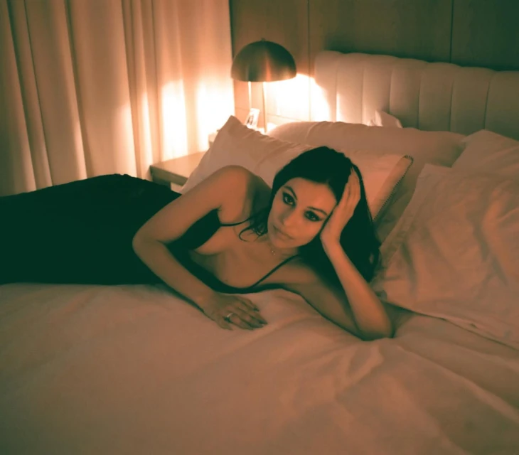 Brunette woman in a black dress lounges on a white bed in a warmly lit bedroom with a bedside table and lamp in the background.