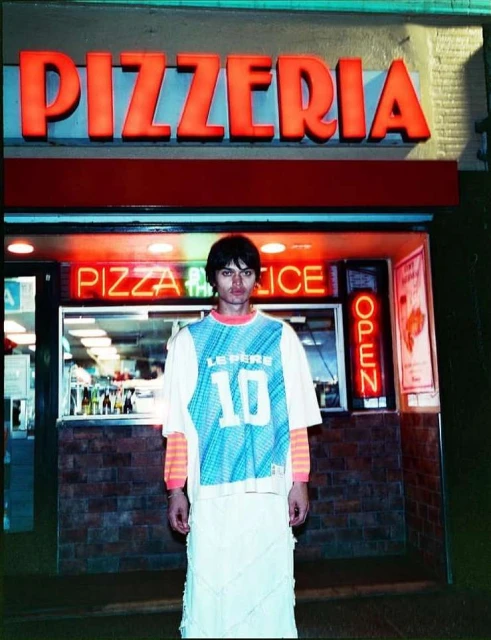Adult stands in front of pizzeria with neon sign. Male model in athletic shirt and white skirt poses in urban setting.
