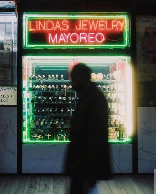 Neon sign illuminates a jewelry store display case filled with watches and cross pendants, with a blurry figure in the foreground.