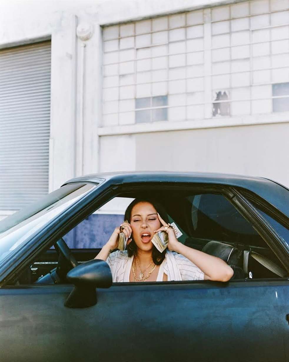 Woman in car holds stacks of money to ears. Blue car, neutral background, light skin, short hair, wearing light colored blouse and jewelry.