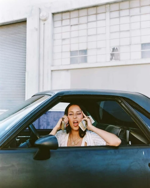 Woman in car holds stacks of money to ears. Blue car, neutral background, light skin, short hair, wearing light colored blouse and jewelry.