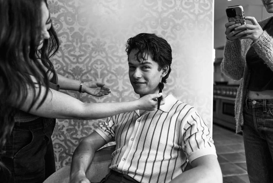 Indoor black and white portrait of a young man getting his hair styled by a hairstylist with another woman standing nearby.