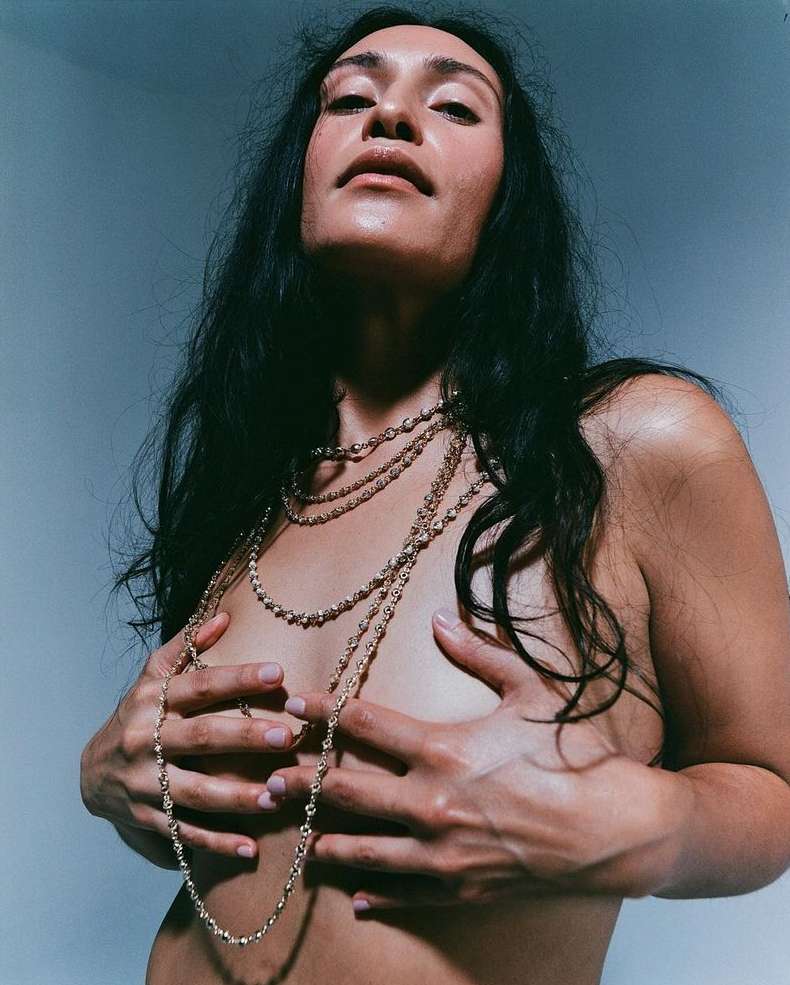 Portrait of a nude adult woman with long dark hair wearing beaded necklaces posing in a studio with soft, neutral lighting and a gray backdrop.