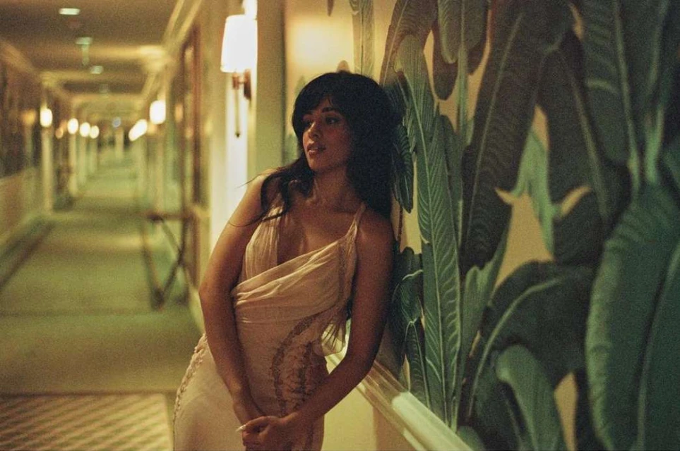 An elegant woman in a light-colored dress poses in a hotel hallway with decorative tropical wallpaper and warm lighting. Interior fashion shot.