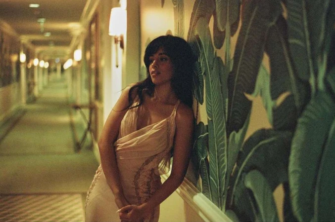 An elegant woman in a light-colored dress poses in a hotel hallway with decorative tropical wallpaper and warm lighting. Interior fashion shot.