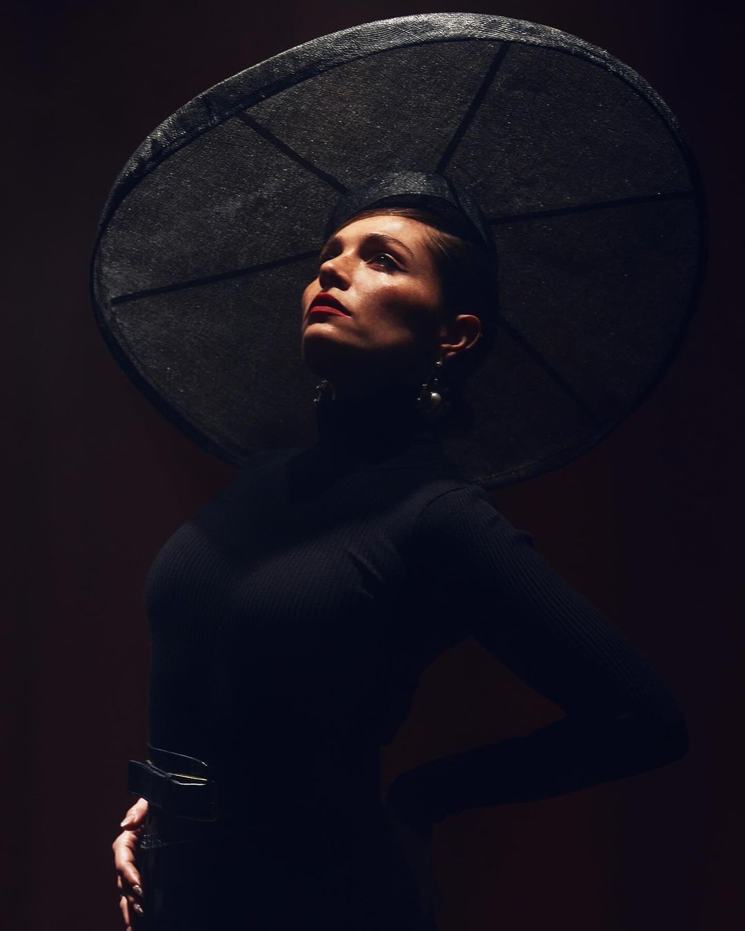 Portrait of a woman with a large hat, wearing a black turtleneck. She is glancing upward in a dramatic pose in a dark studio.