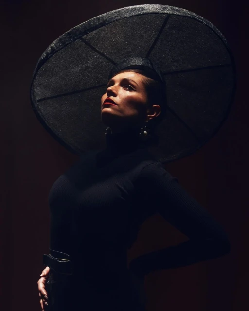 Portrait of a woman with a large hat, wearing a black turtleneck. She is glancing upward in a dramatic pose in a dark studio.