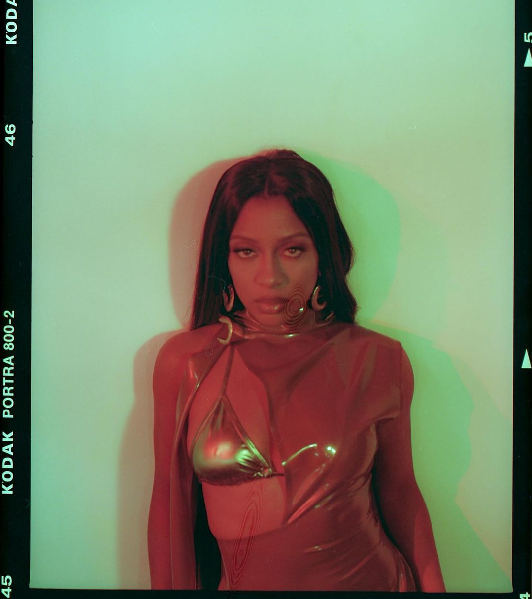 Woman in a metallic dress with gold jewelry posing in front of a green and white wall in a portrait photograph with studio lighting.