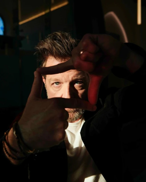 Adult man frames face with hands in indoor portrait. Man has fair skin, facial hair, and wears bracelets, a t-shirt, and a jacket.