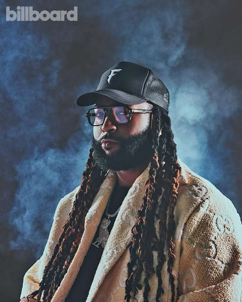 Portrait of adult male wearing glasses, cap, and patterned jacket in a studio setting with a smoky background, featuring the Billboard logo.
