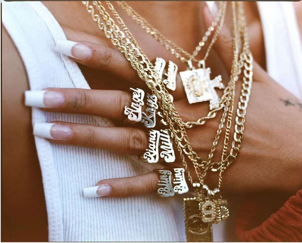 Close-up on hand wearing custom name rings, accessorized with multiple layered gold chains and pendants against a white tank top.