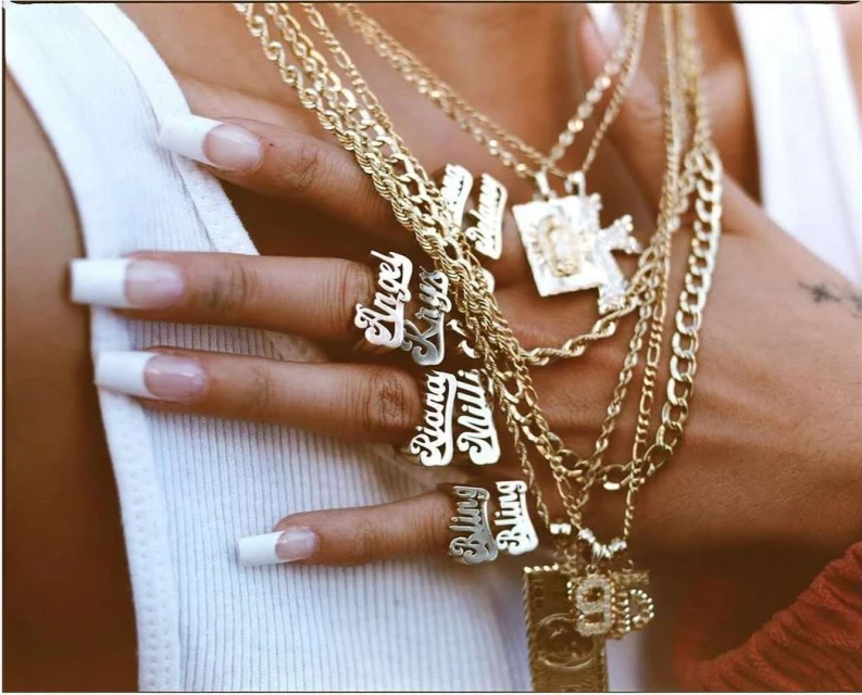 Close-up on hand wearing custom name rings, accessorized with multiple layered gold chains and pendants against a white tank top.