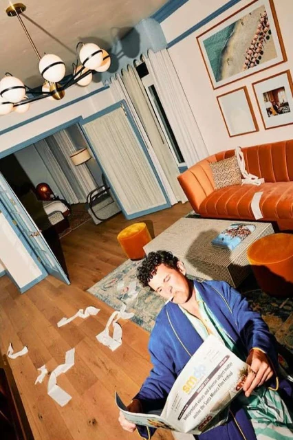 Man in robe reads newspaper in messy interior living room with orange sofa, carpet, artwork, scattered paper, and modern lighting fixture.