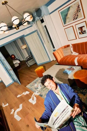 Man in robe reads newspaper in messy interior living room with orange sofa, carpet, artwork, scattered paper, and modern lighting fixture.