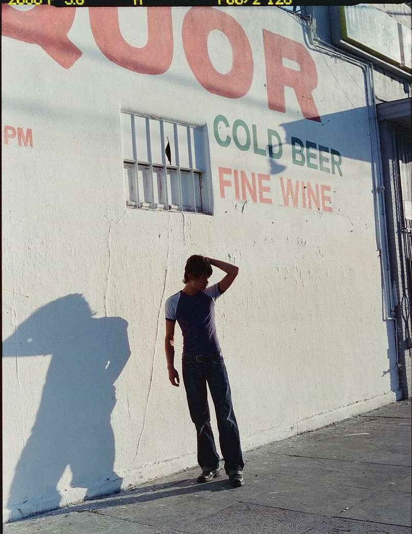 Young man in casual clothing poses against a wall with 'Liquor, Cold Beer, Fine Wine' painted on it, with a shadow alongside him.