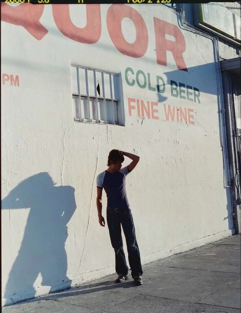 Young man in casual clothing poses against a wall with 'Liquor, Cold Beer, Fine Wine' painted on it, with a shadow alongside him.