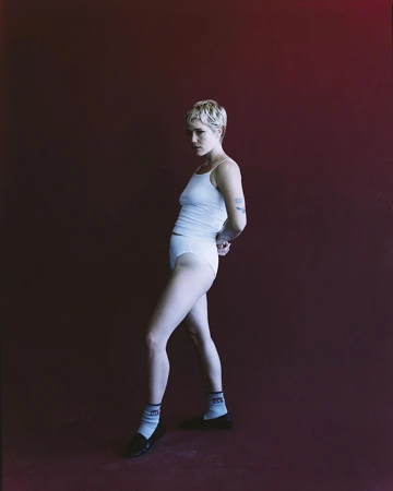 Woman with short blonde hair wearing a white tank top, white briefs, socks, and dark shoes against a dark red backdrop.