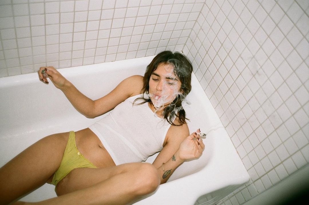 Woman lounges in a white bathtub, smoking. She wears a white top and yellow bottoms, with a white tiled wall in the background.