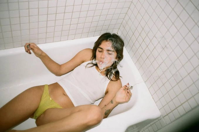 Woman lounges in a white bathtub, smoking. She wears a white top and yellow bottoms, with a white tiled wall in the background.