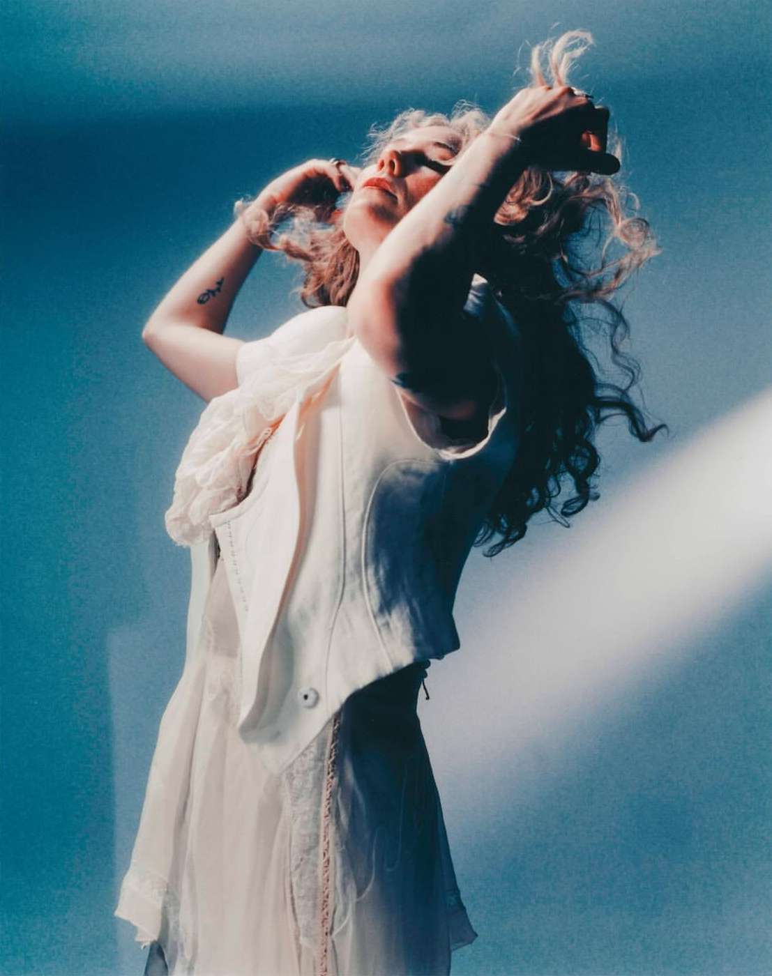 Elegant woman in flowing white dress poses with arms raised against a blue background, a shaft of light adding dimension to the scene.