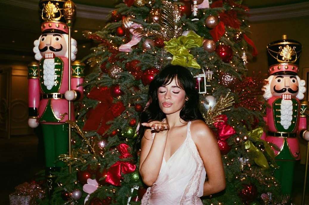 Adult woman poses in front of a Christmas tree, wearing a light-pink dress and blowing a kiss, with decorative nutcrackers visible.