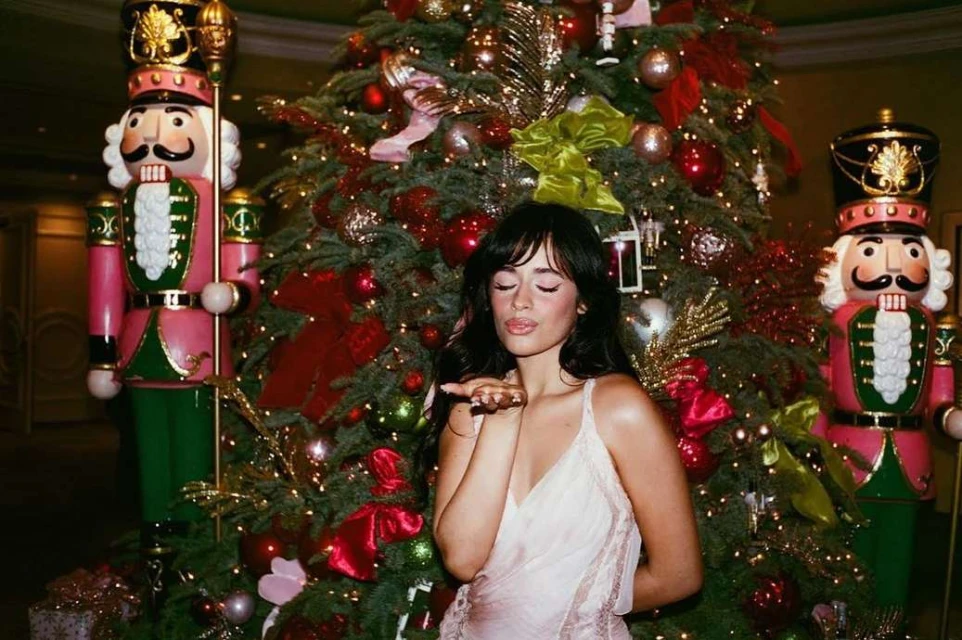 Adult woman poses in front of a Christmas tree, wearing a light-pink dress and blowing a kiss, with decorative nutcrackers visible.