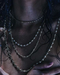 Close-up of layered silver chain necklaces on a person's chest with dark wet hair under moody blue lighting.