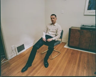 Man with a mustache seated on a green chair wearing a cream button-down shirt and black pants in a wood-floored room.