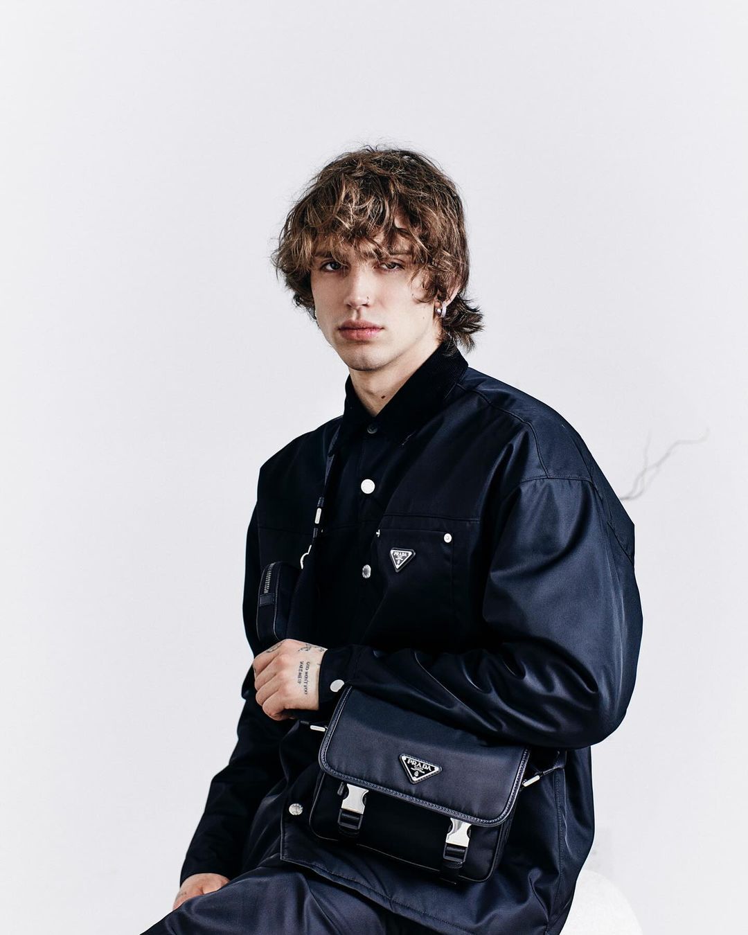 Portrait of a young adult male with curly hair, wearing a dark jacket and crossbody bag against a white background in a studio setting.