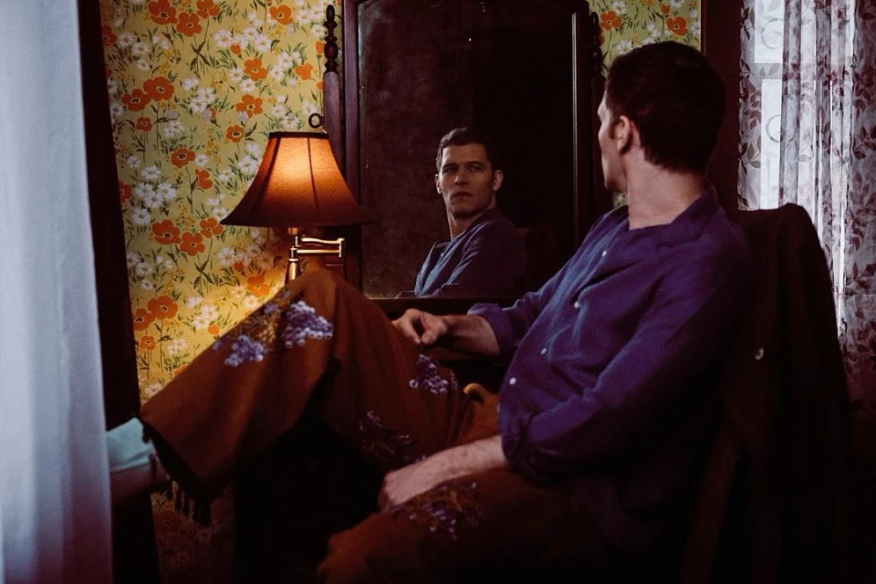 Man in patterned pajamas and purple shirt sits in chair, reflected in dark mirror with floral wallpaper in background indoors.