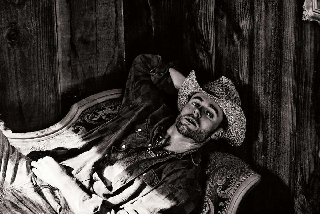 Monochrome portrait of a man in a cowboy hat reclining on an ornate chaise lounge against a rustic wood-paneled wall backdrop.