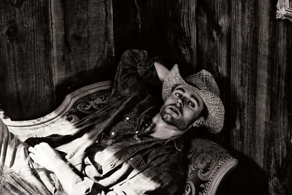 Monochrome portrait of a man in a cowboy hat reclining on an ornate chaise lounge against a rustic wood-paneled wall backdrop.