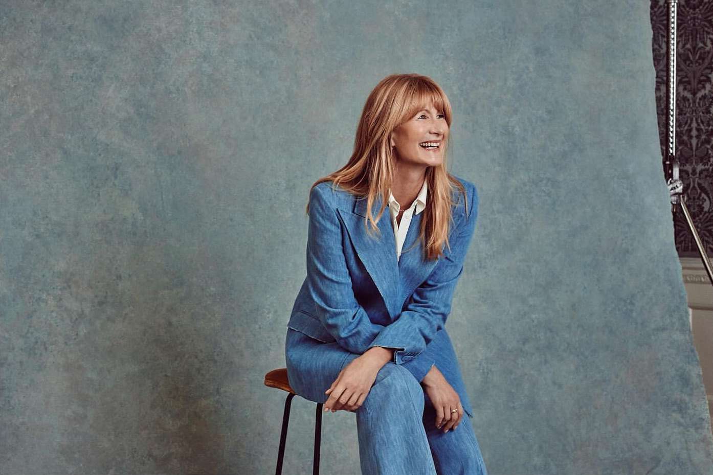 Smiling woman with long blonde hair in a blue suit sitting on a stool in a photo studio with a blue textured background.
