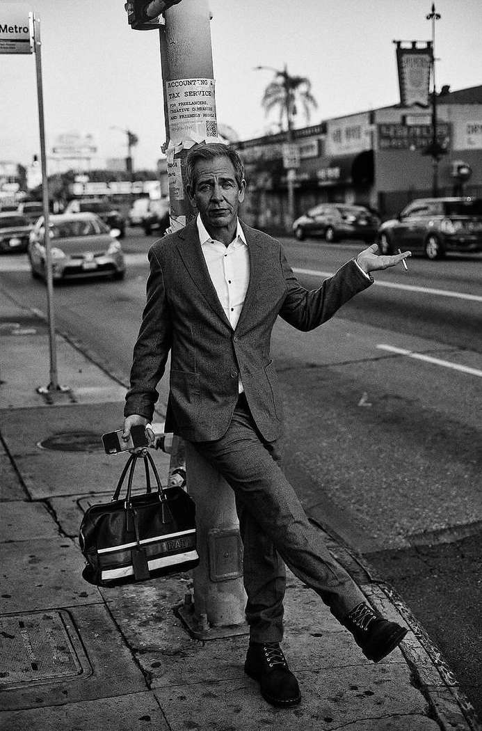 Man in suit poses on urban sidewalk holding a bag, with street traffic and buildings in the background in a black and white photograph.